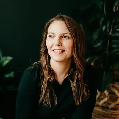 A woman with long brown hair smiles while looking to the side. She is wearing a black top, with a dark, leafy background.