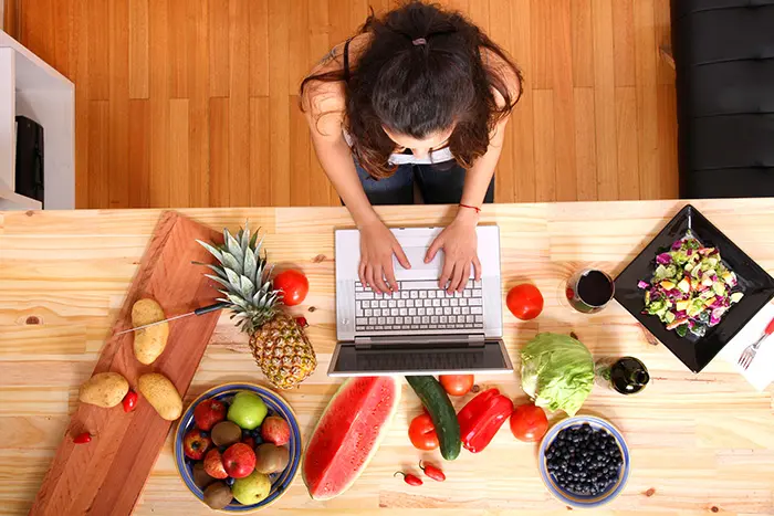 Overhead view of a person typing on a laptop at a wooden table surrounded by fresh fruits and vegetables, including a pineapple, watermelon, and salad, illustrating a healthy lifestyle.