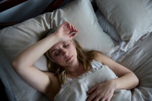 A woman lies in bed looking tired or unwell, with one hand resting on her forehead and a neutral expression on her face. She is covered by a beige blanket and surrounded by light-colored pillows.