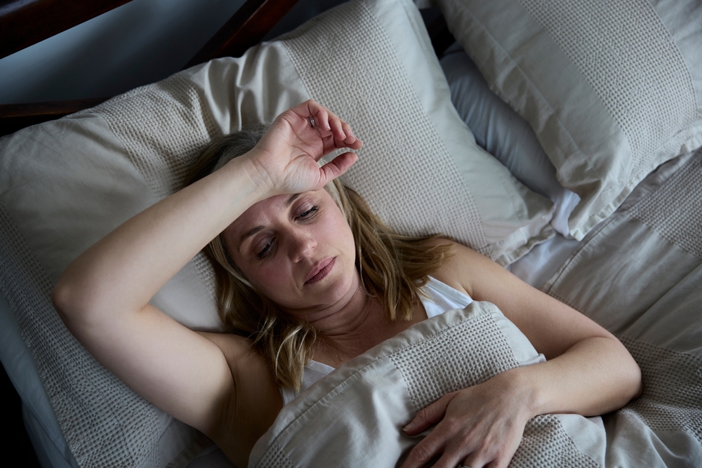 A woman lies in bed looking tired or unwell, with one hand resting on her forehead and a neutral expression on her face. She is covered by a beige blanket and surrounded by light-colored pillows.